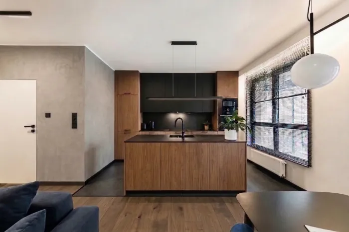 The same apartment after the renovation: dark graphite tiles in the kitchen and smoked oak planks in the living area, the left wall in warm grey microcement, wood island fronts and a slim black linear pendant over the island