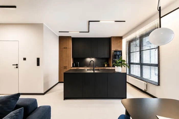 A living room with an open kitchen: a light floor, a black zigzag ceiling light, a light wall with a white door on the left, a black kitchen island in front of wood tall units and black wall cabinets, and a large window with blinds on the right